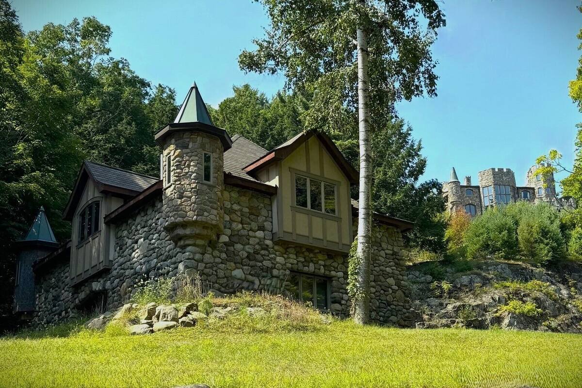 A stone house with a turret and a large window, surrounded by greenery and a castle in the background.