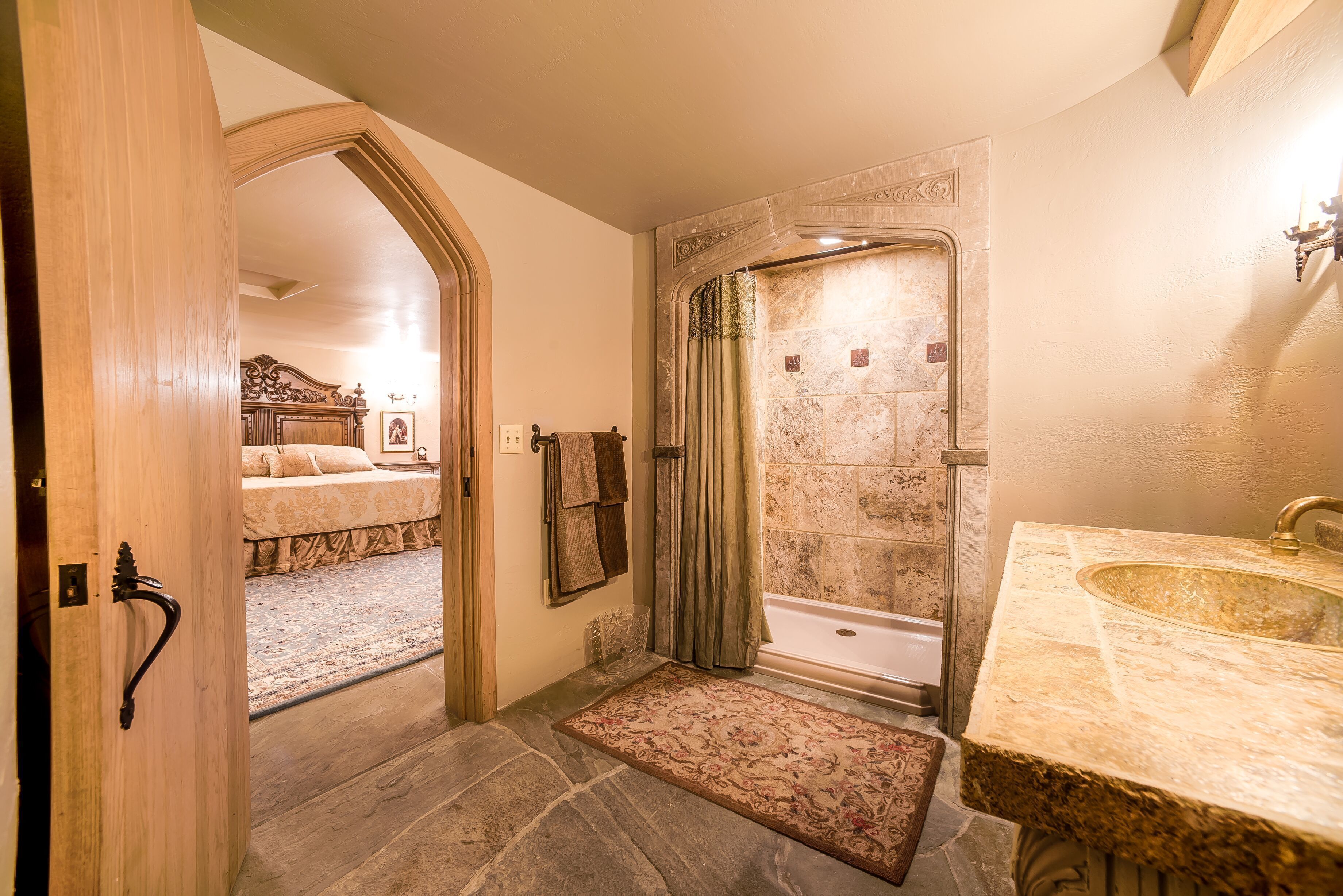 Ein Badezimmer mit Steinboden, einer Badewanne, einem Waschbecken und einer Tür zu einem Schlafzimmer mit Bett und Teppich.