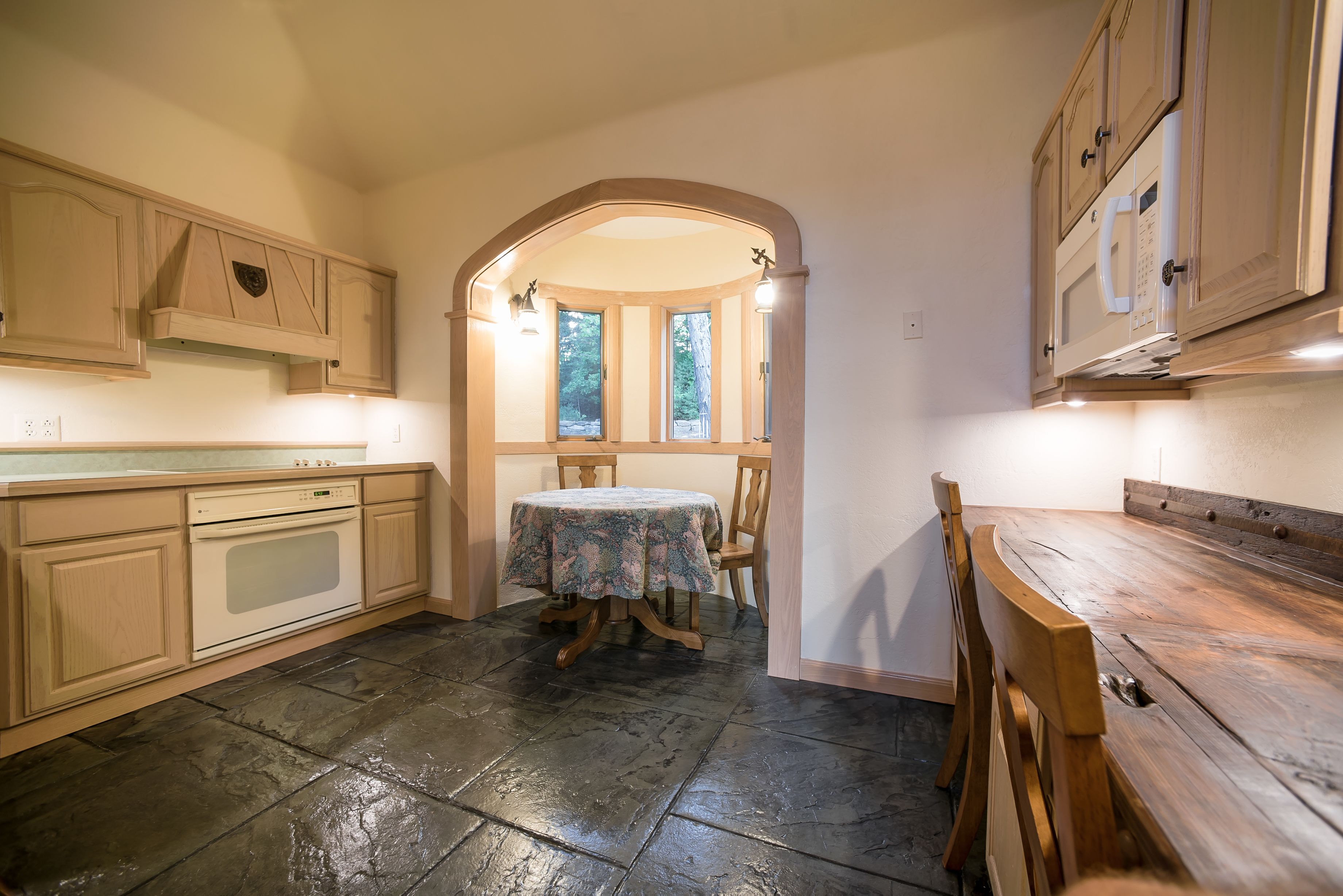 Een keuken met stenen vloer, houten kastjes, een eettafel met stoelen en uitzicht door een raam.