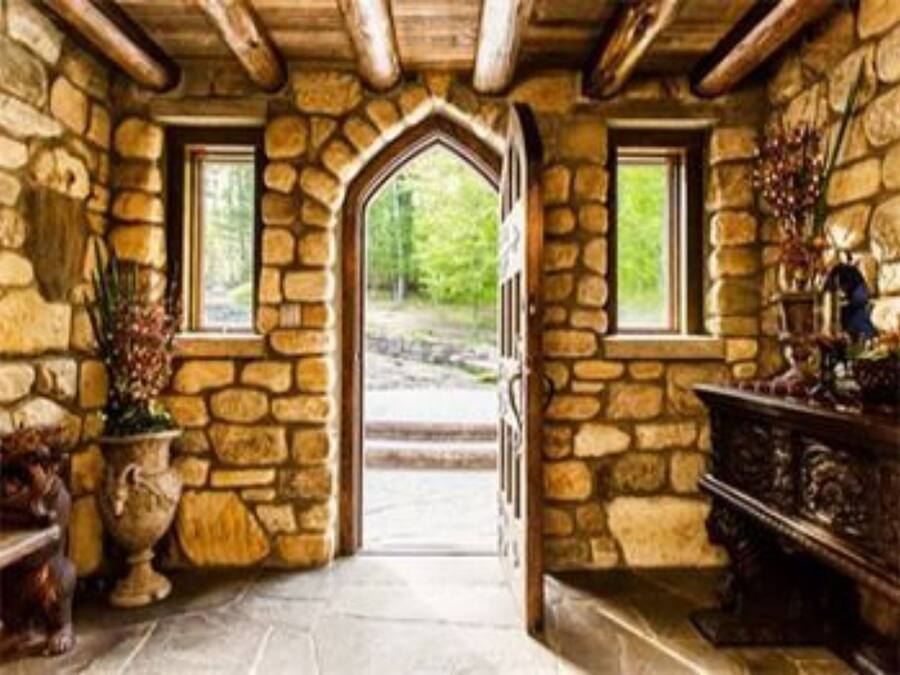 A stone-walled room with a wooden ceiling, a large arched doorway, and a view of a forested area outside.