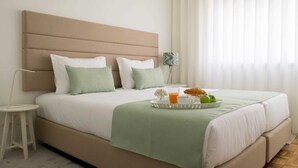 Uma cama bem arrumada com um cobertor verde, um tabuleiro com frutas e um copo de sumo de laranja, e um abajur.