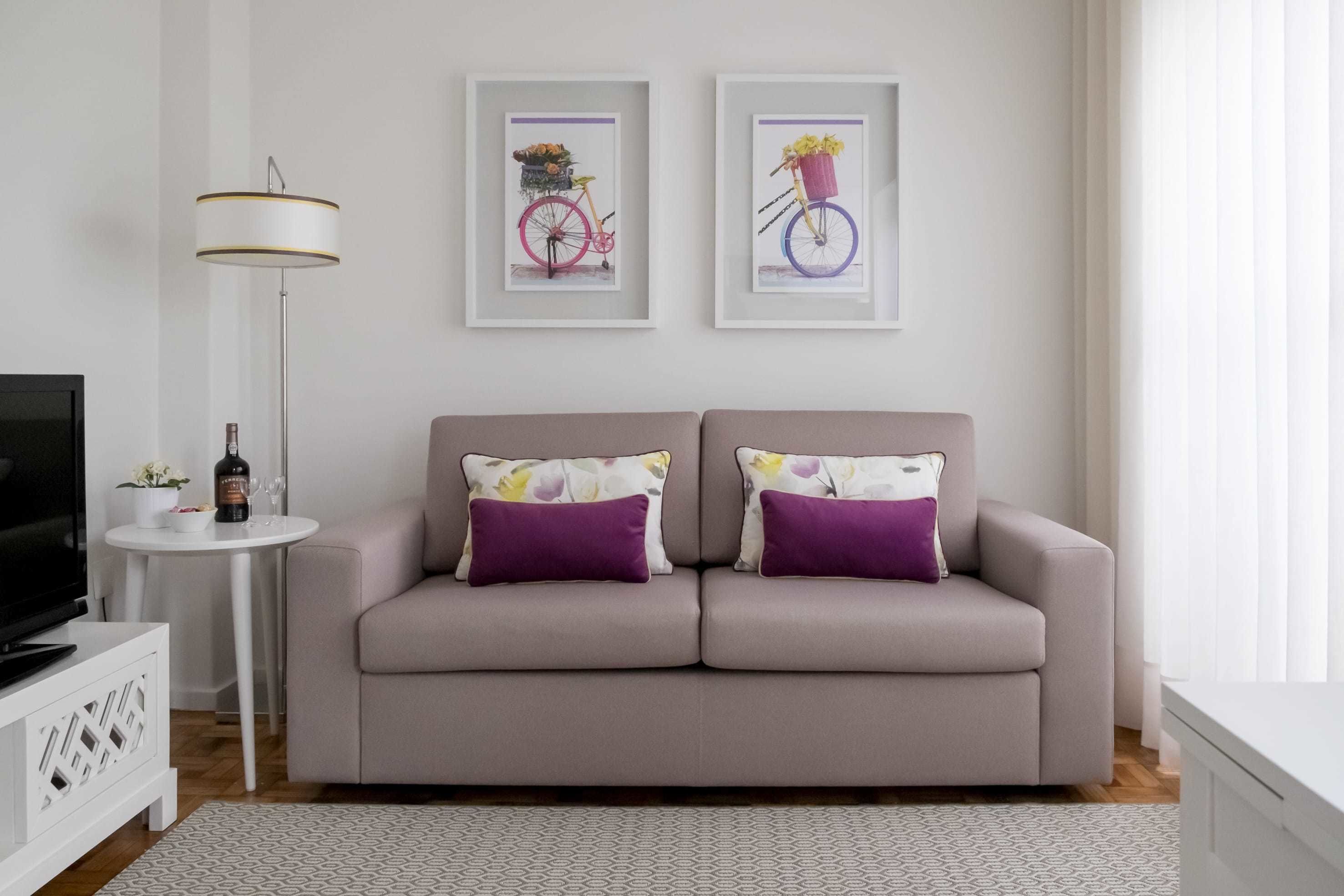 A living room with a beige sofa, two purple cushions, a small white side table with a bottle and glasses, a floor lamp, and two framed artworks on the wall.