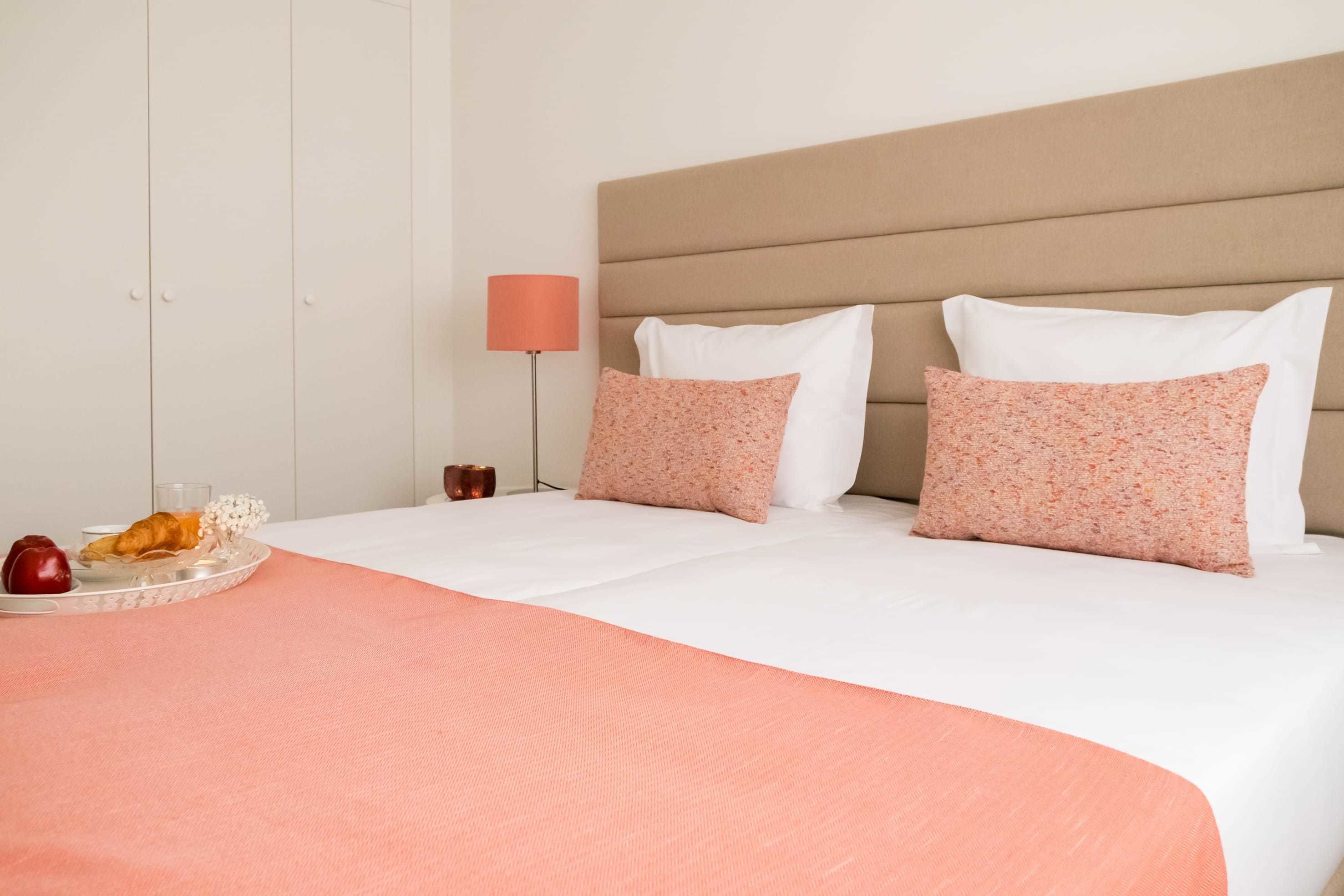 Habitación de hotel con cama, dos almohadas, mesita de noche con lámpara y bandeja con comida.