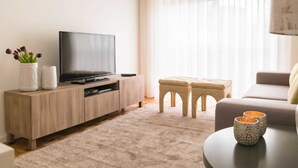 Uma sala de estar moderna com uma televisão de tela plana em um armário de madeira, um tapete bege, um banquinho estampado e uma pequena mesa com xícaras decorativas.
