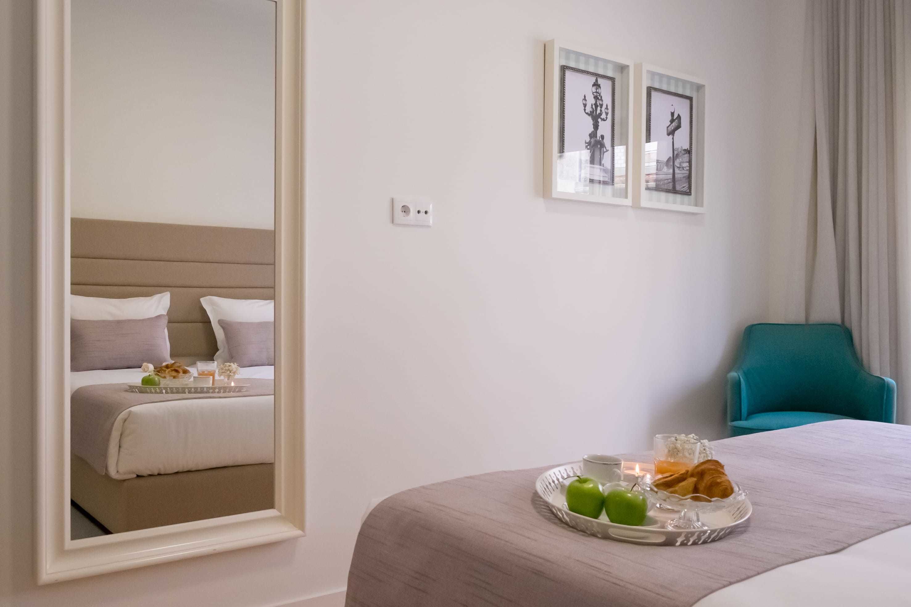 A hotel room with a bed, a bedside table with breakfast items, a chair, and framed pictures on the wall.