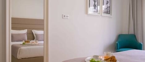 A hotel room with a bed, a bedside table with breakfast items, a chair, and framed pictures on the wall.