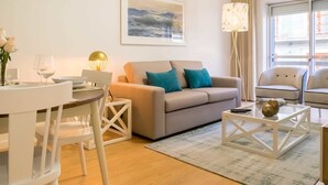 Uma sala de estar moderna com um sofá bege, almofadas verde-água, uma mesa de centro branca e uma mesa de vidro com uma taça de vinho e um vaso.