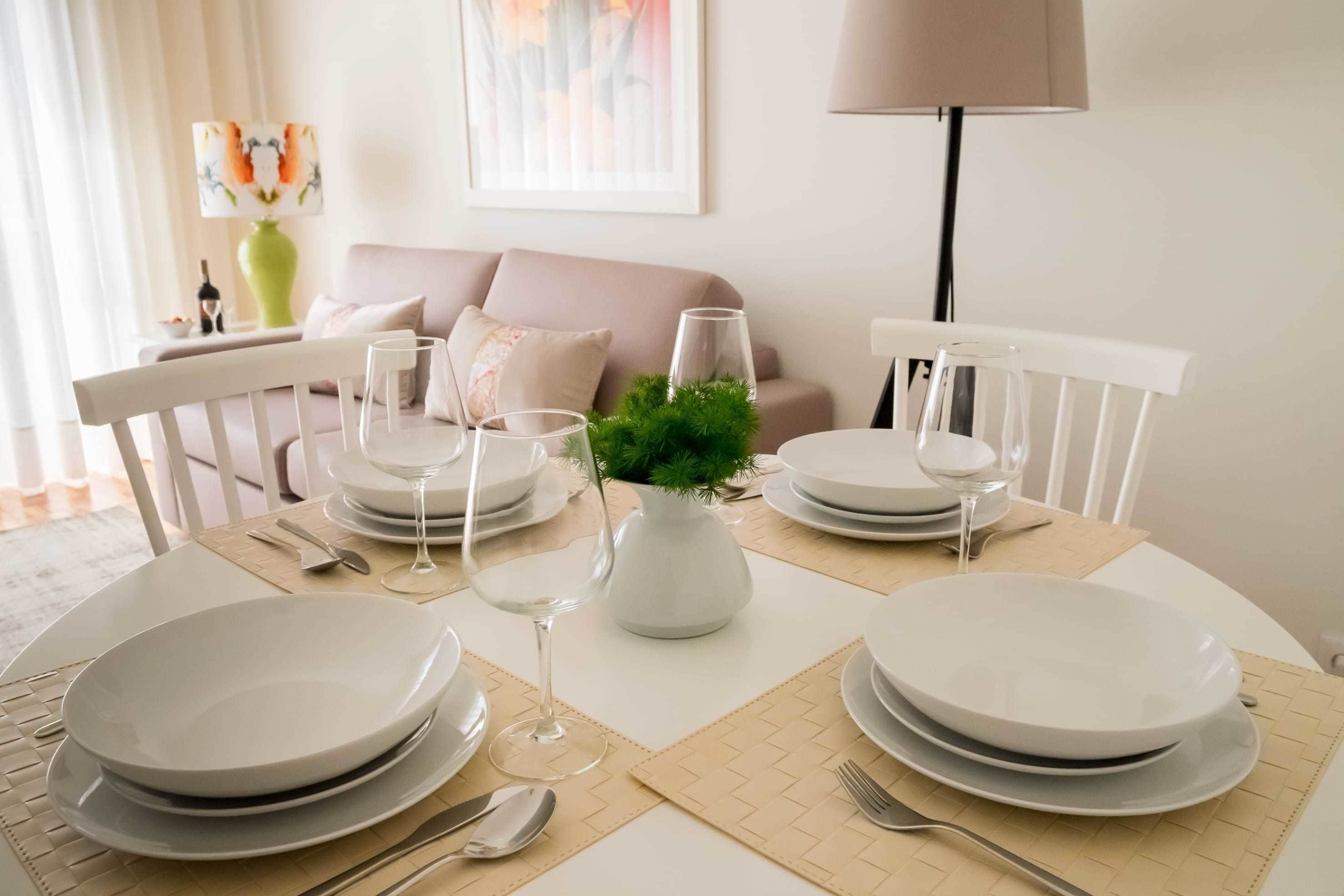 Une table à manger dressée avec de l’assiette blanche, des verres et des couverts, un vase rempli de feuillage vert et une lampe dans un salon moderne.