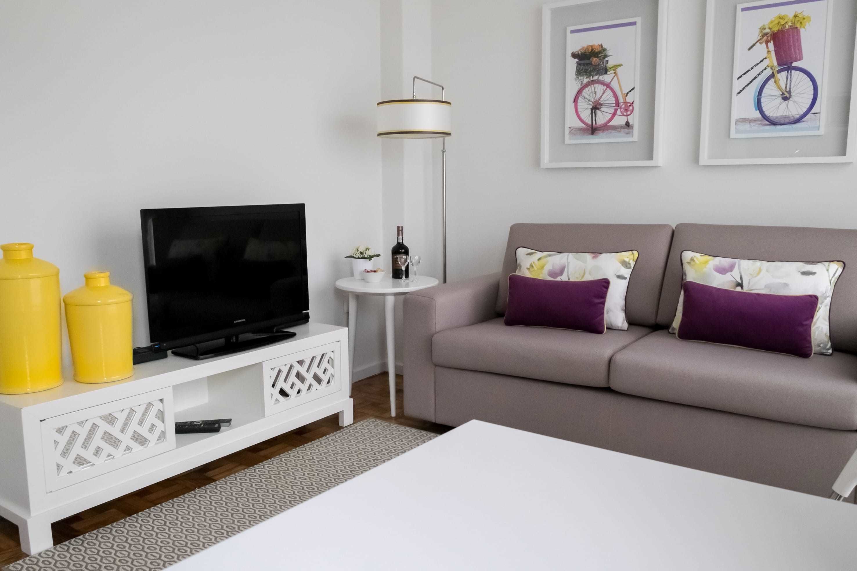 Un salon moderne avec un téléviseur à écran plat, une petite table d’appoint blanche sur laquelle se trouvent une bouteille et un verre, un canapé gris agrémenté de coussins violets, et deux tableaux encadrés accrochés au mur.