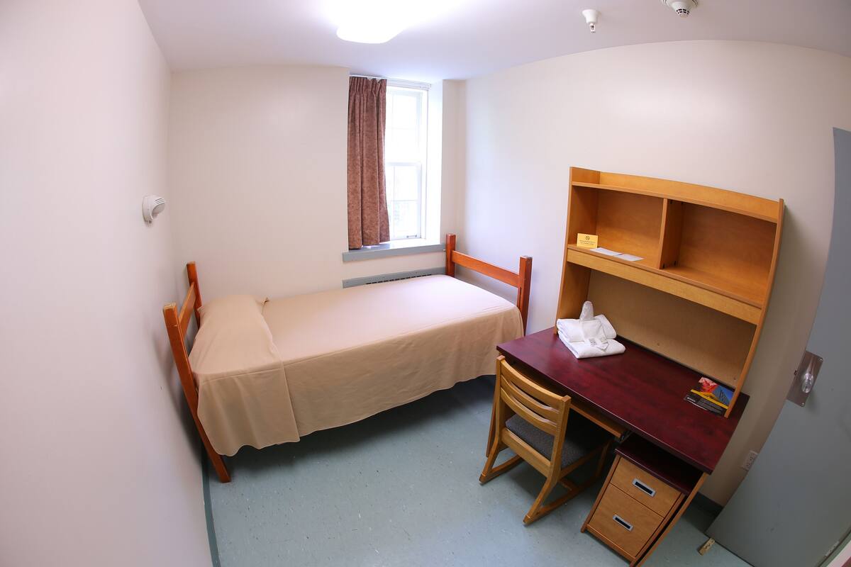 A small, single-bed room with a wooden desk, chair, and a shelf.