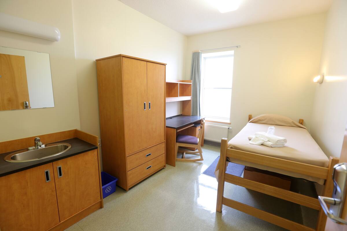 A small, clean room with a wooden bed, a desk, a chair, a wardrobe, and a bathroom with a sink and mirror.