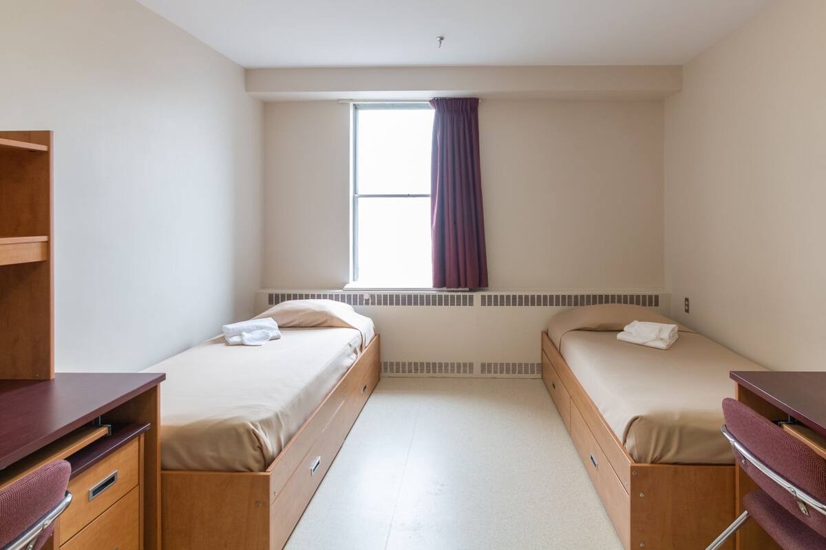 A compact room with two beds, wooden desks, and a window with curtains.