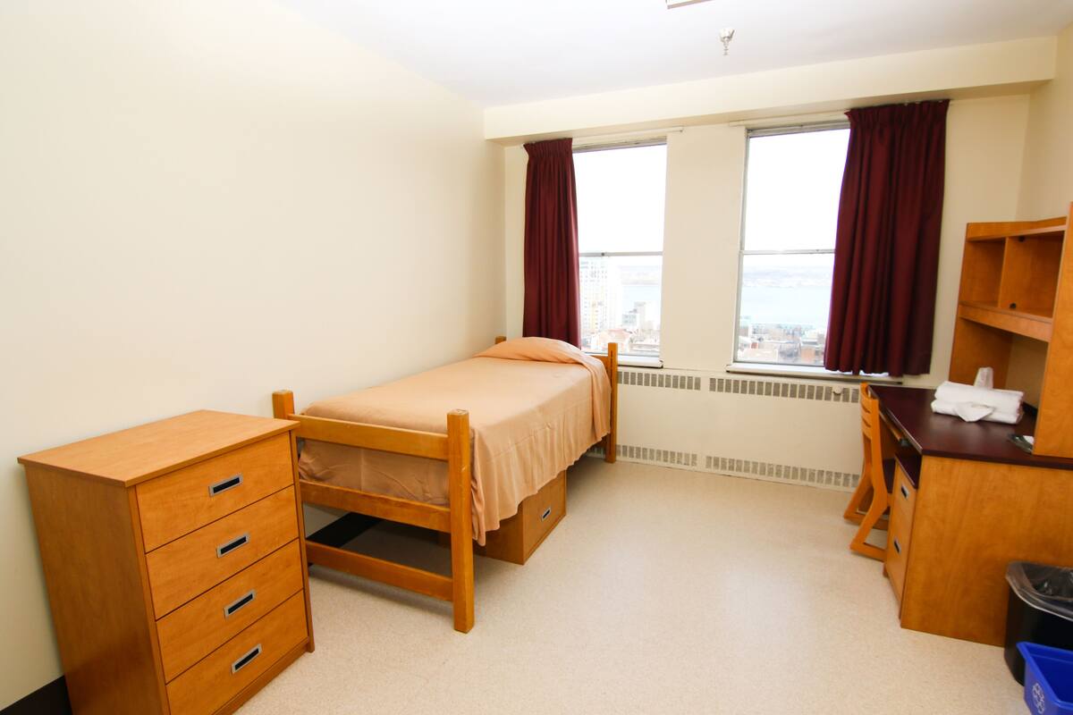 A bedroom with a single bed, a wooden dresser, a desk, and a window with curtains.