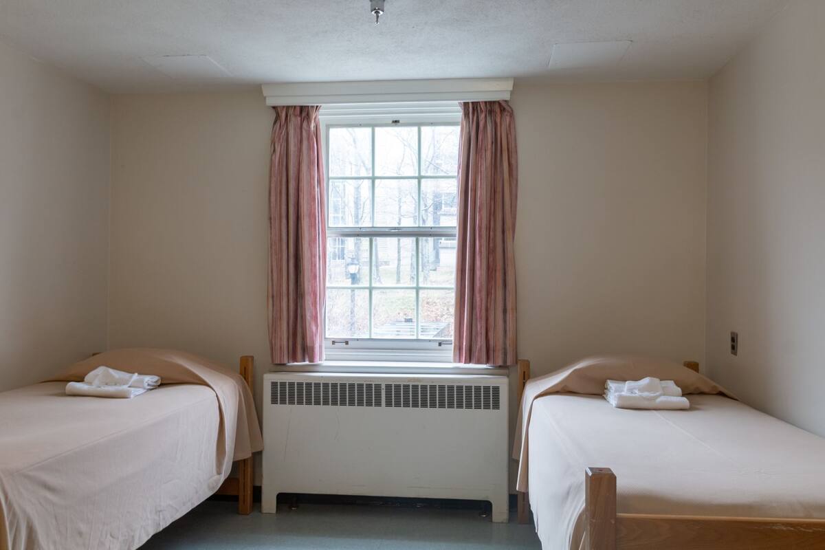 A room with two single beds, a window with curtains, a radiator, and a view of the outside.