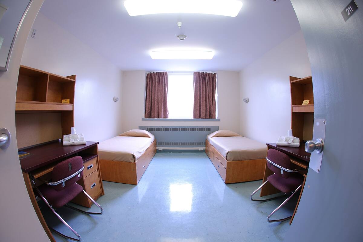 A small, well-lit room with two beds, a desk, and a chair.