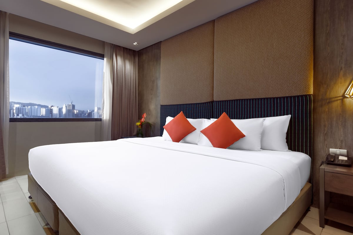 A large bed with white bedding and orange pillows, a wooden headboard, a nightstand with a phone, and a view of a cityscape through a large window.