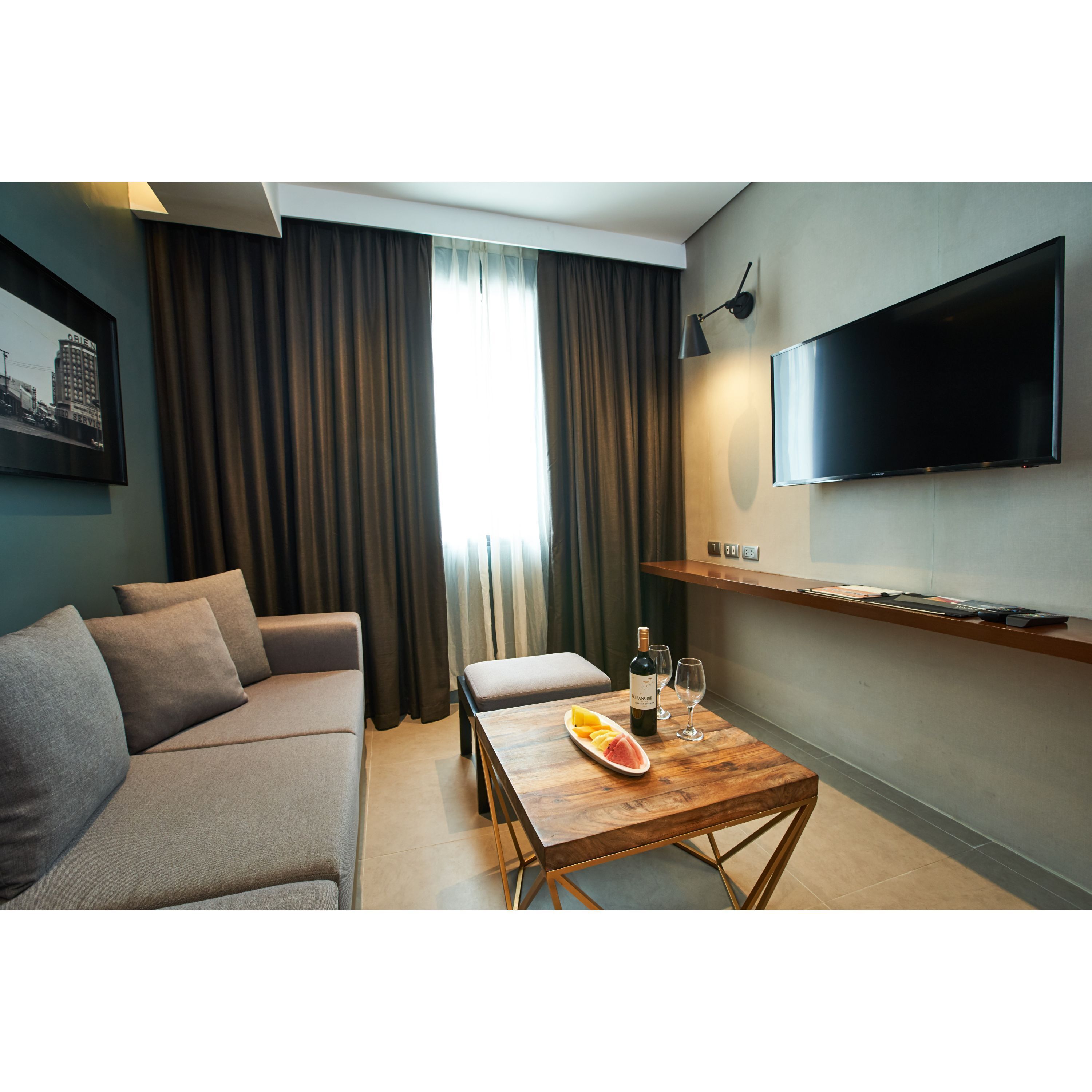 A modern hotel room with a sofa, a wooden coffee table with a bottle of wine and glasses, a flat-screen TV mounted on the wall, and a cityscape picture on the wall.