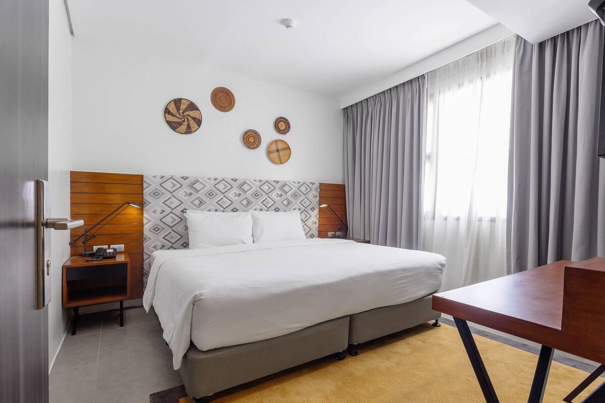 A modern hotel room with a large bed, a wooden headboard, a desk, and a view of the outdoors through a window with curtains.