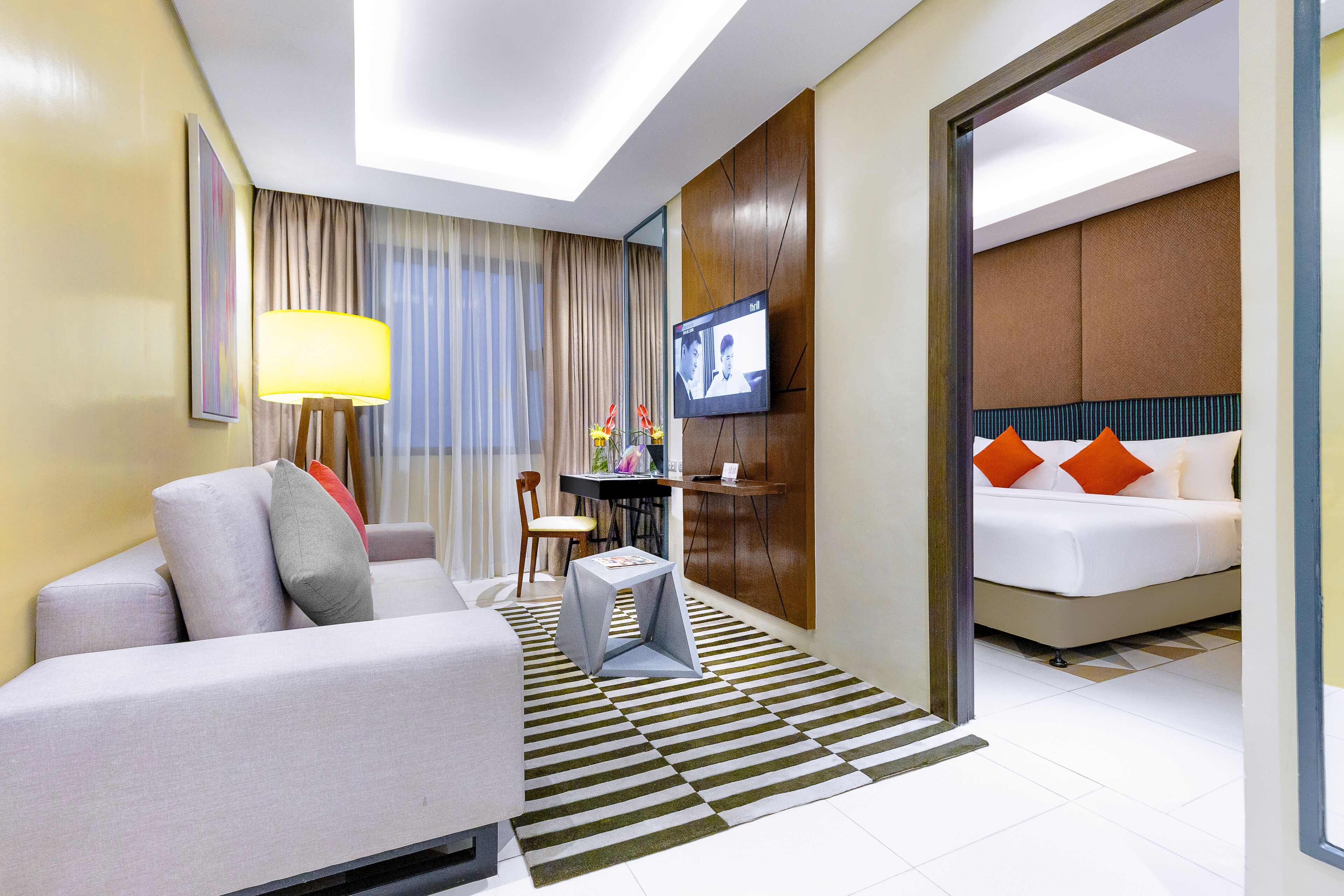 A hotel room with a sofa, a small table, a TV, and a bed with orange pillows.