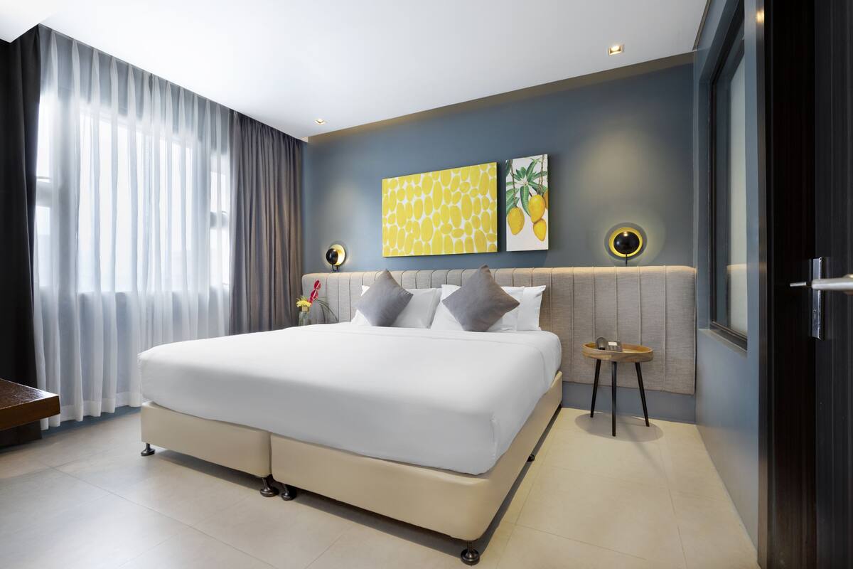 A modern hotel room with a large bed, a bedside table, a small round table, and a wall-mounted light fixture.