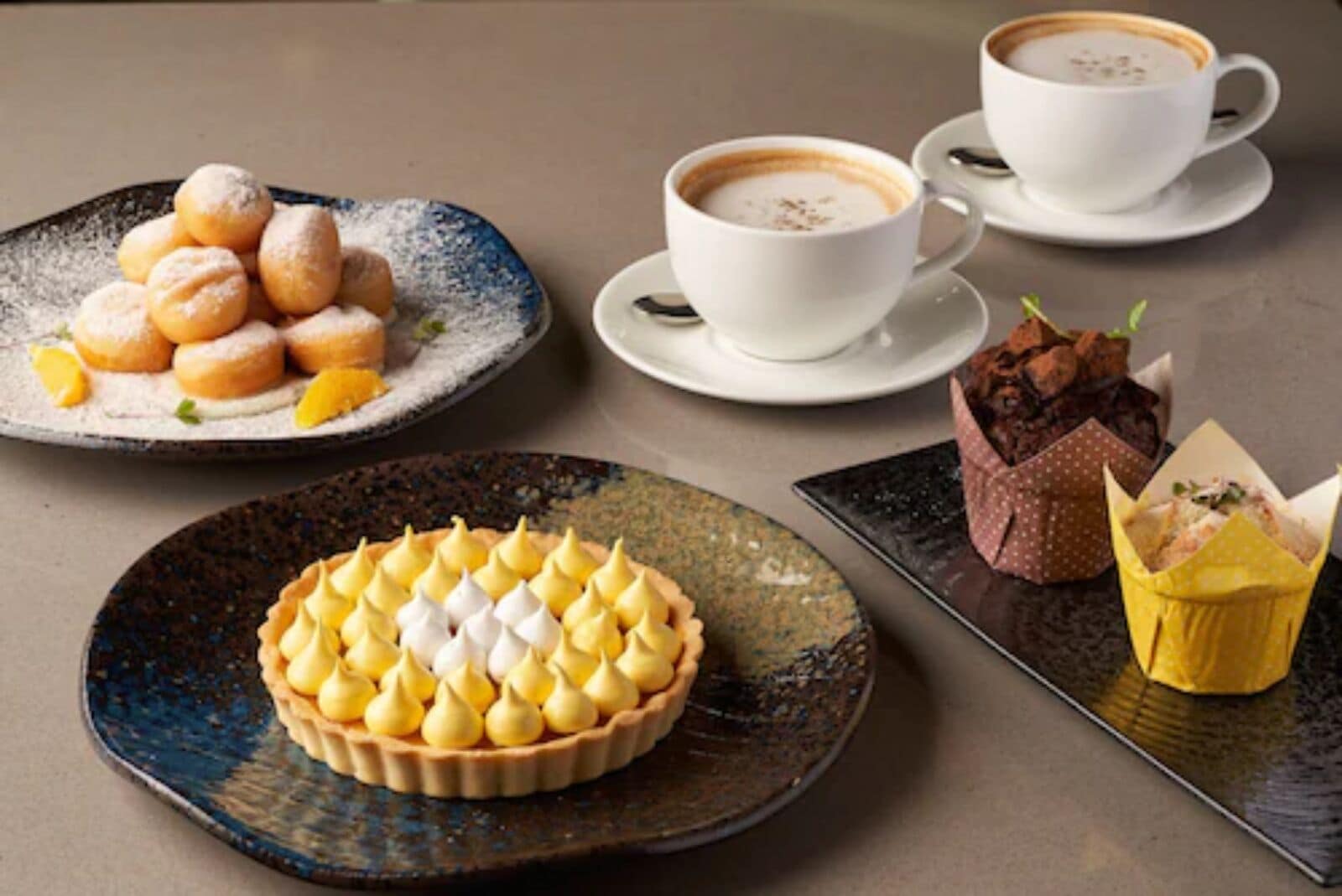 A table with a dessert plate of lemon meringue pie, a plate of powdered sugar-covered pastries, two cups of coffee, and a chocolate dessert in a paper cup.