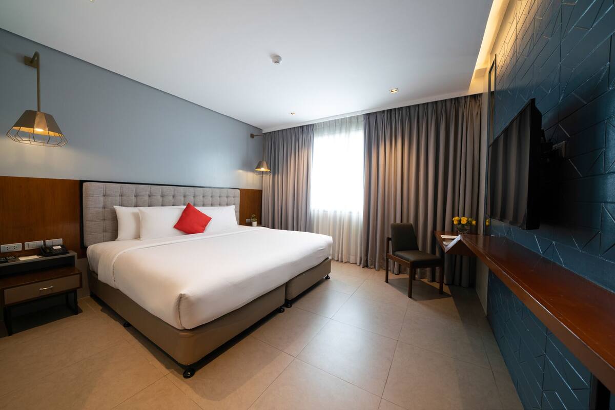 A hotel room with a large bed, a desk with a chair, a TV mounted on the wall, and a window with curtains.