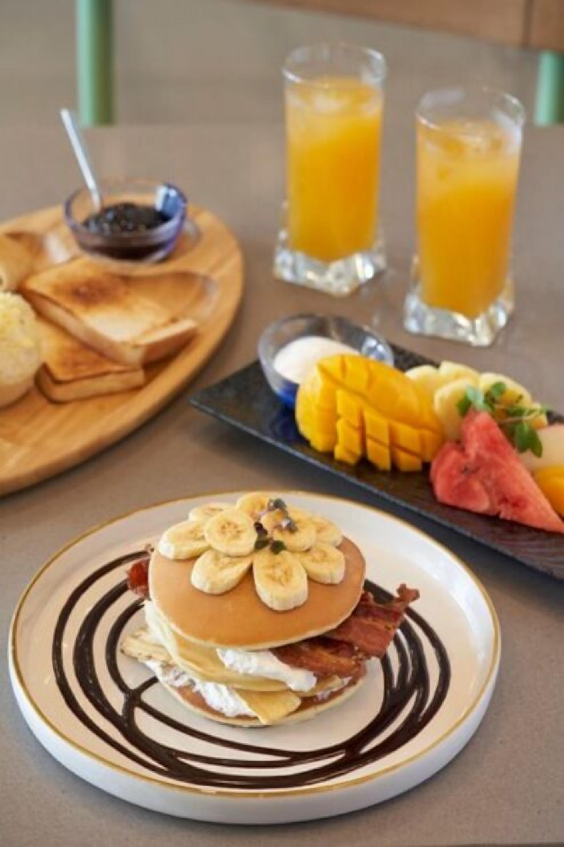 A breakfast setting with a plate of pancakes topped with bananas and bacon, a plate of sliced fruits, and two glasses of orange juice.