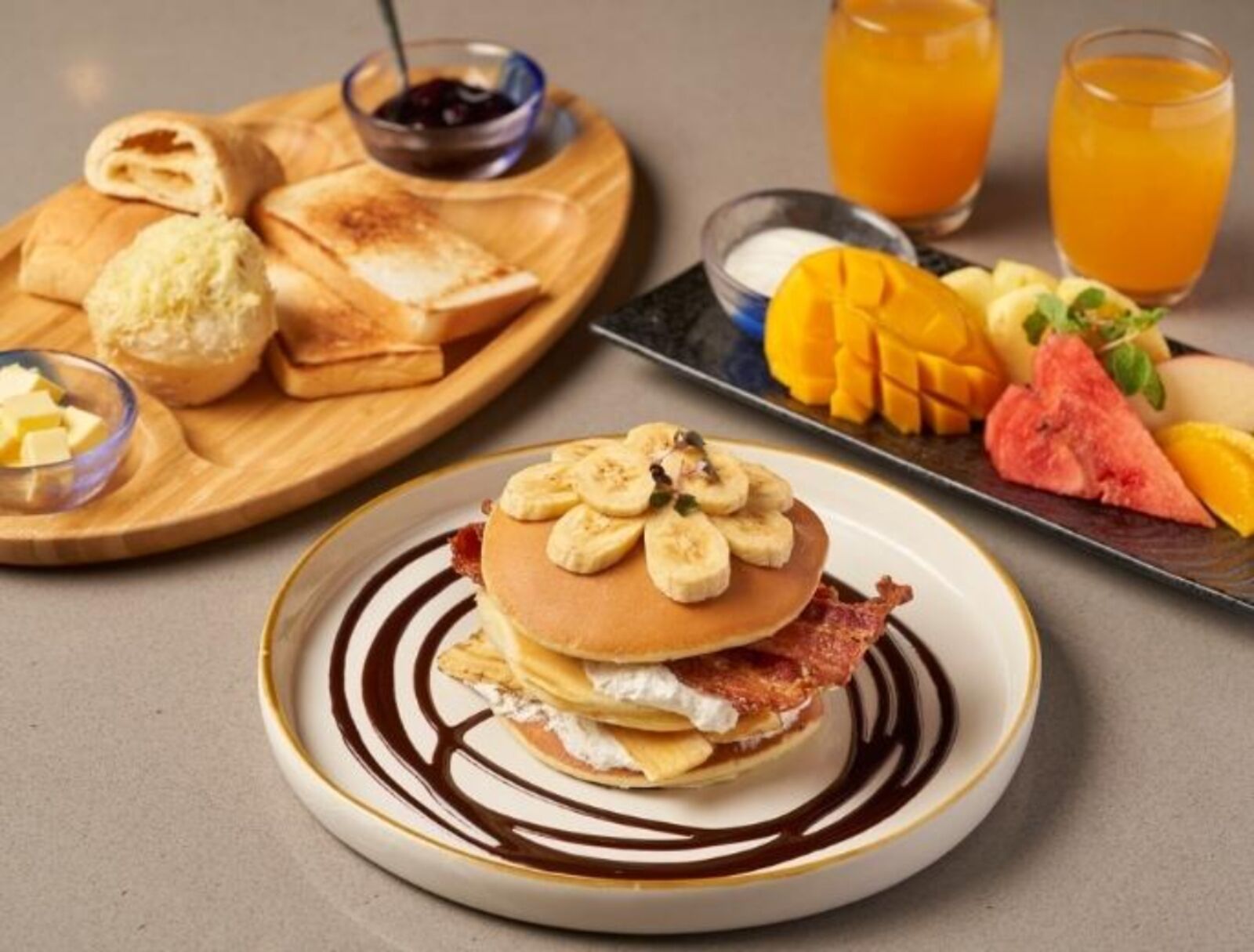 Ein Frühstücksteller mit Pfannkuchen, Speck und Bananen, eine Holzplatte mit Toast und Marmelade sowie ein Obstteller mit Mango und Wassermelone.