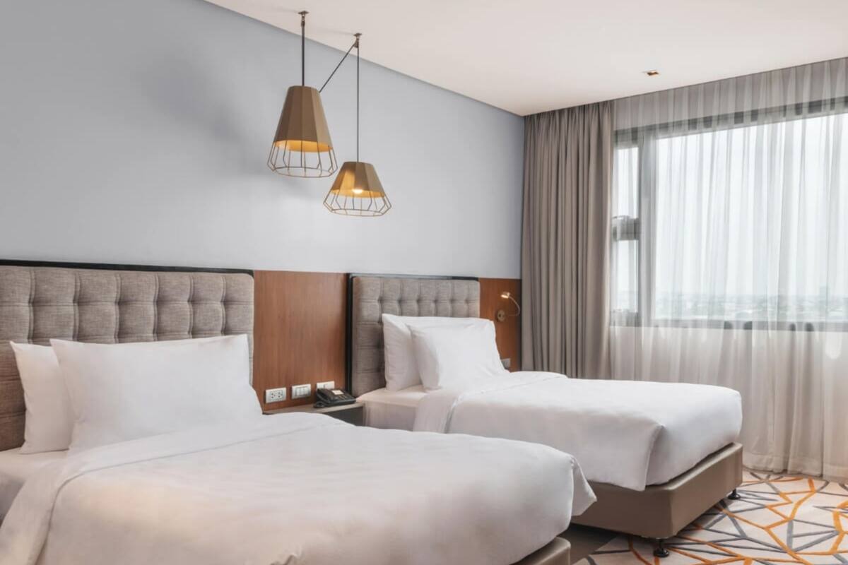 A hotel room with two beds, a wooden headboard, a nightstand, a telephone, and a window with curtains.