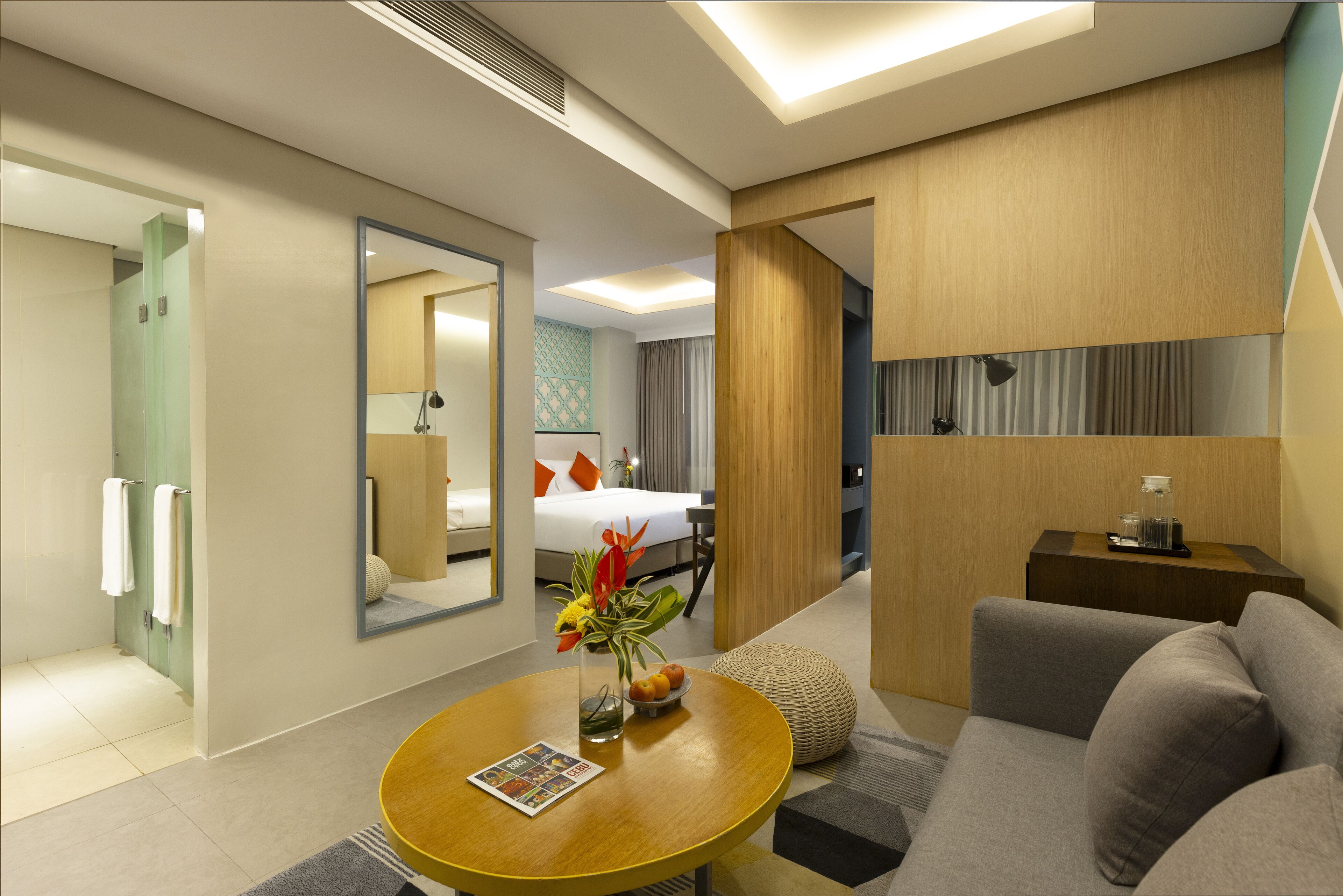 A modern hotel room with a sofa, a round wooden table with a magazine, a mirror, and a bed with a white duvet.