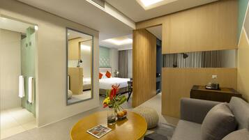 A modern hotel room with a sofa, a round wooden table with a magazine, a mirror, and a bed with a white duvet.