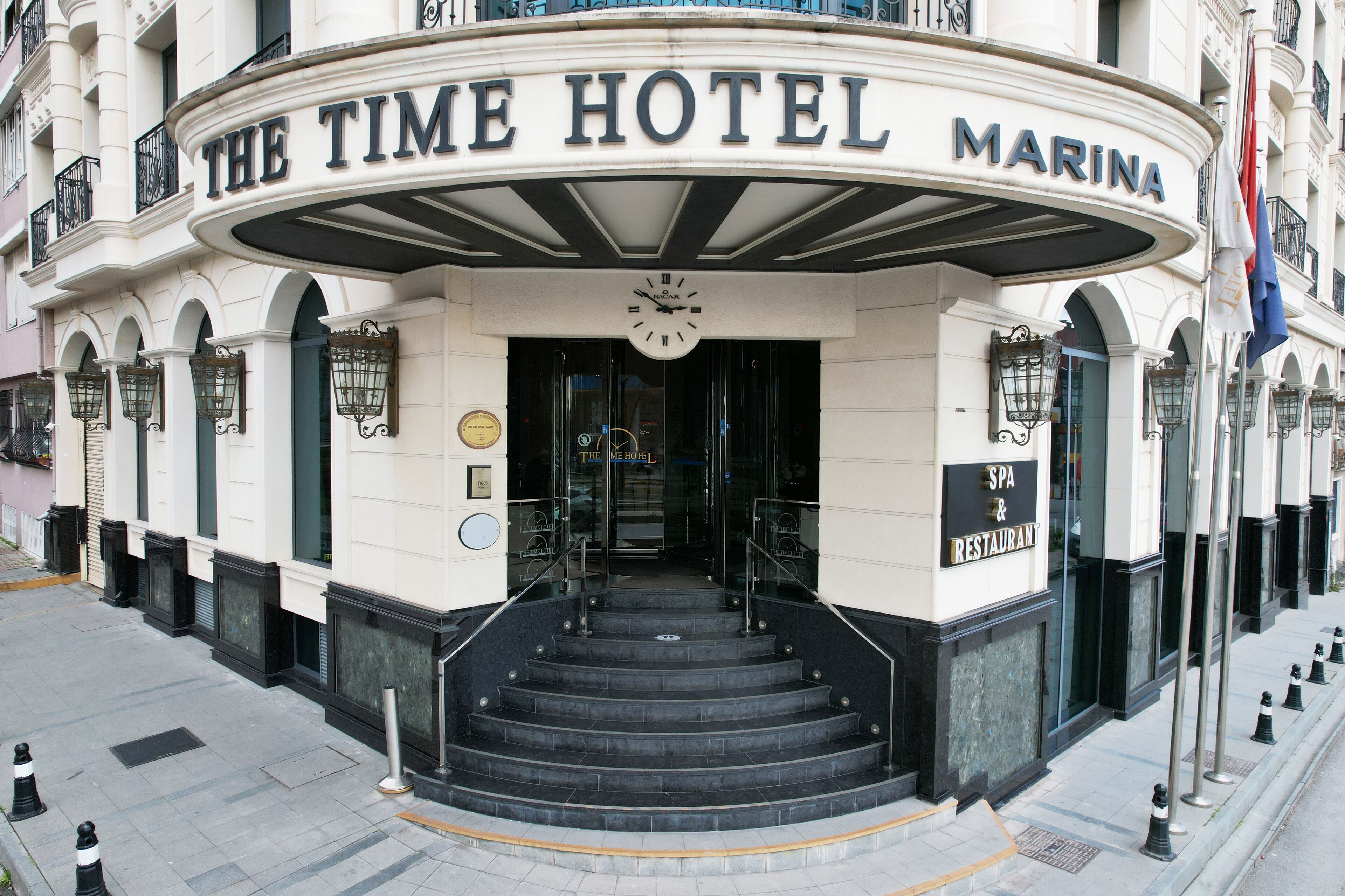 Entrada do Time Hotel Marina, com um relógio acima da porta, um letreiro para o spa e restaurante, e um conjunto de degraus que levam à entrada.