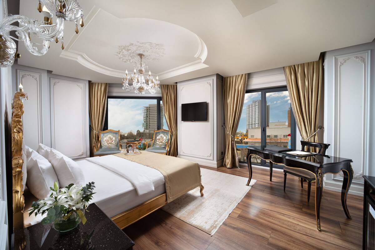 A luxurious bedroom with a large bed, a chandelier, a desk, and a view of the city.