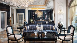 Un hall d’entrée luxueux avec un sol en marbre, un imposant lustre, un canapé bleu moelleux agrémenté de coussins blancs, et un bar classique en arrière-plan.