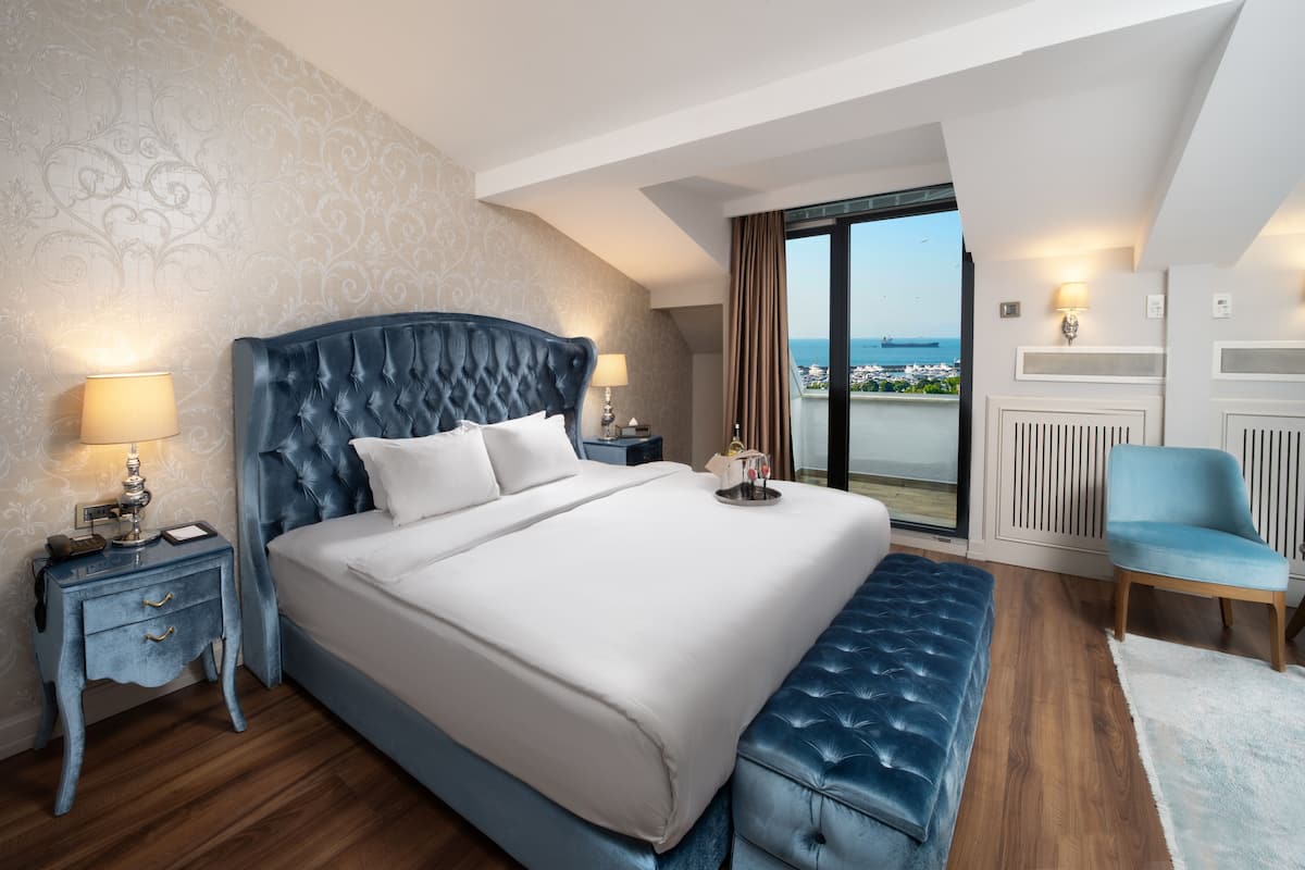 A hotel room with a large bed, a nightstand, a chair, and a view of the sea.