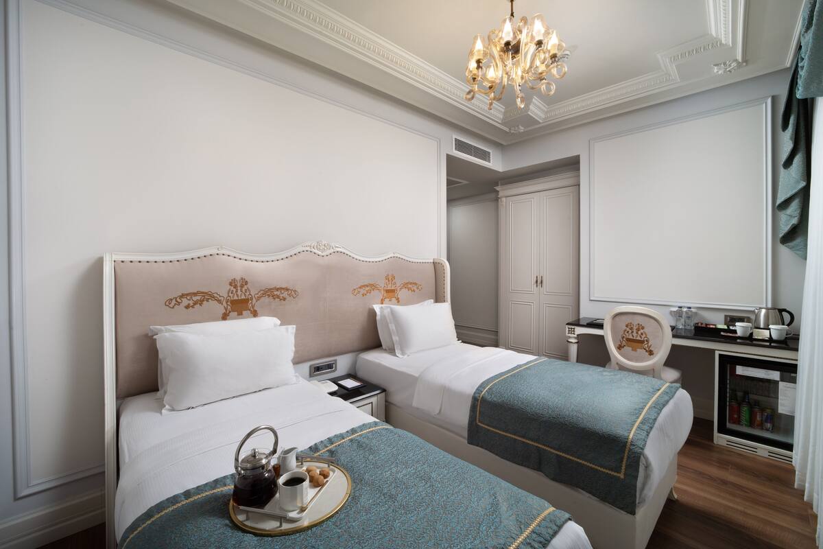 A hotel room with two beds, a desk, a chair, a minibar, and a chandelier.