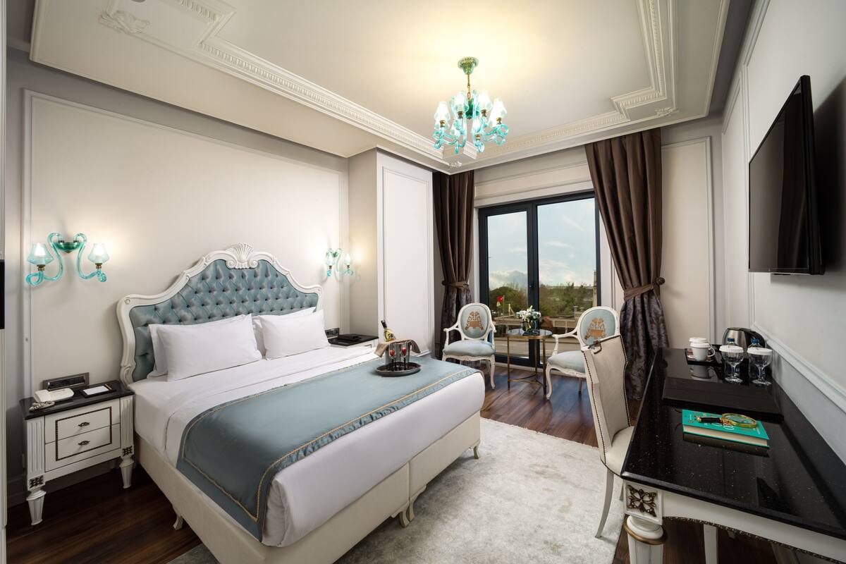 A bedroom with a large bed, a desk, a chair, a chandelier, and a television.