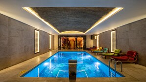 Une piscine intérieure au design moderne, entourée de chaises longues et dotée d’un plafond haut avec un système d’éclairage.