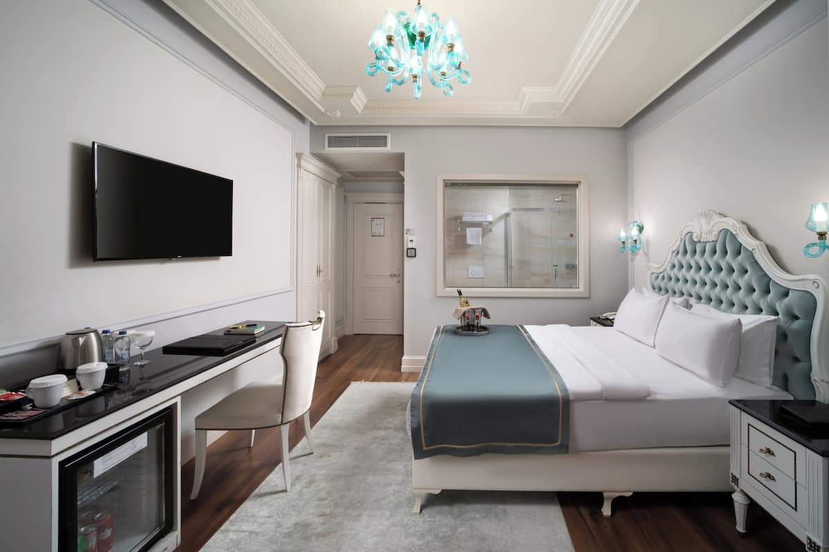 Een moderne hotelkamer met een flatscreen televisie, een bureau met een koffieapparaat, een bed met blauwe accenten en een kroonluchter.