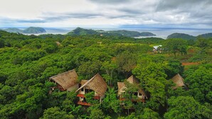 Un paysage tropical luxuriant, avec plusieurs charmantes chaumières nichées au milieu d’une végétation dense, dominant une zone côtière avec des collines lointaines et un ciel nuageux.
