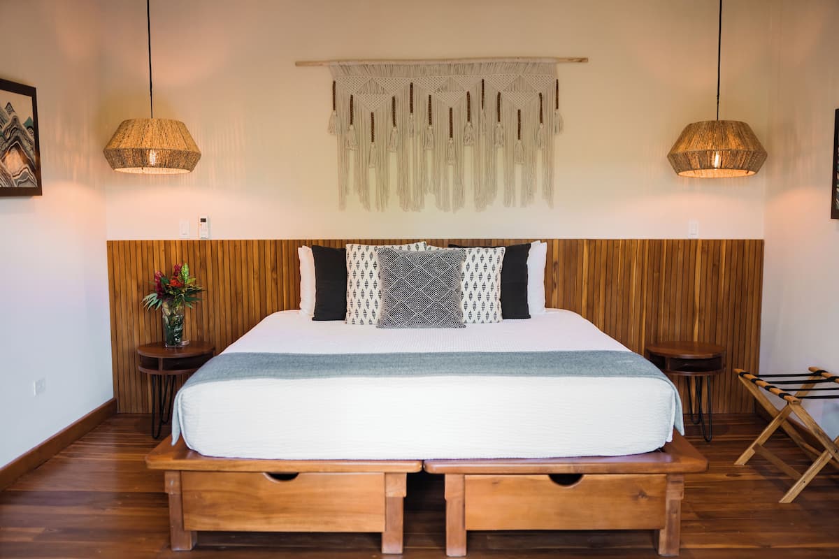 A bedroom with a wooden headboard, a large bed with patterned pillows, two bedside tables, and a hanging macrame piece above the bed.
