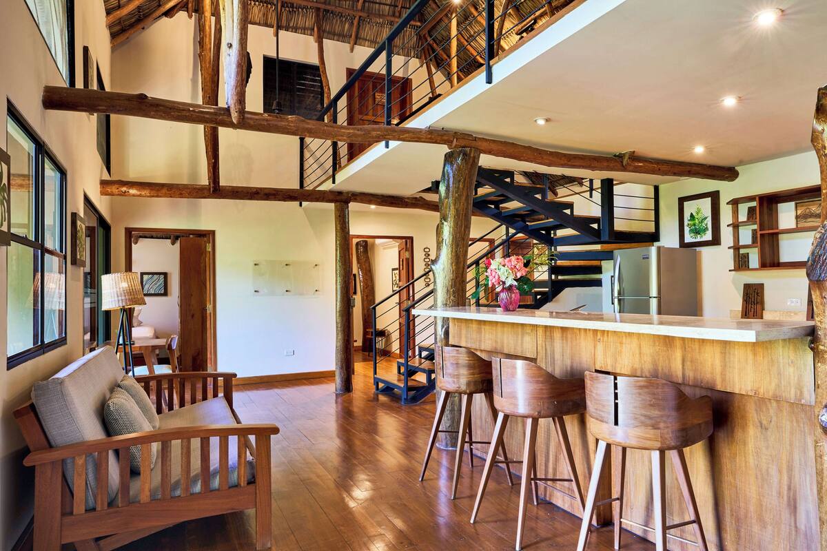 A spacious living area with a wooden staircase, a bar area with stools, and a seating area with a sofa and chairs.