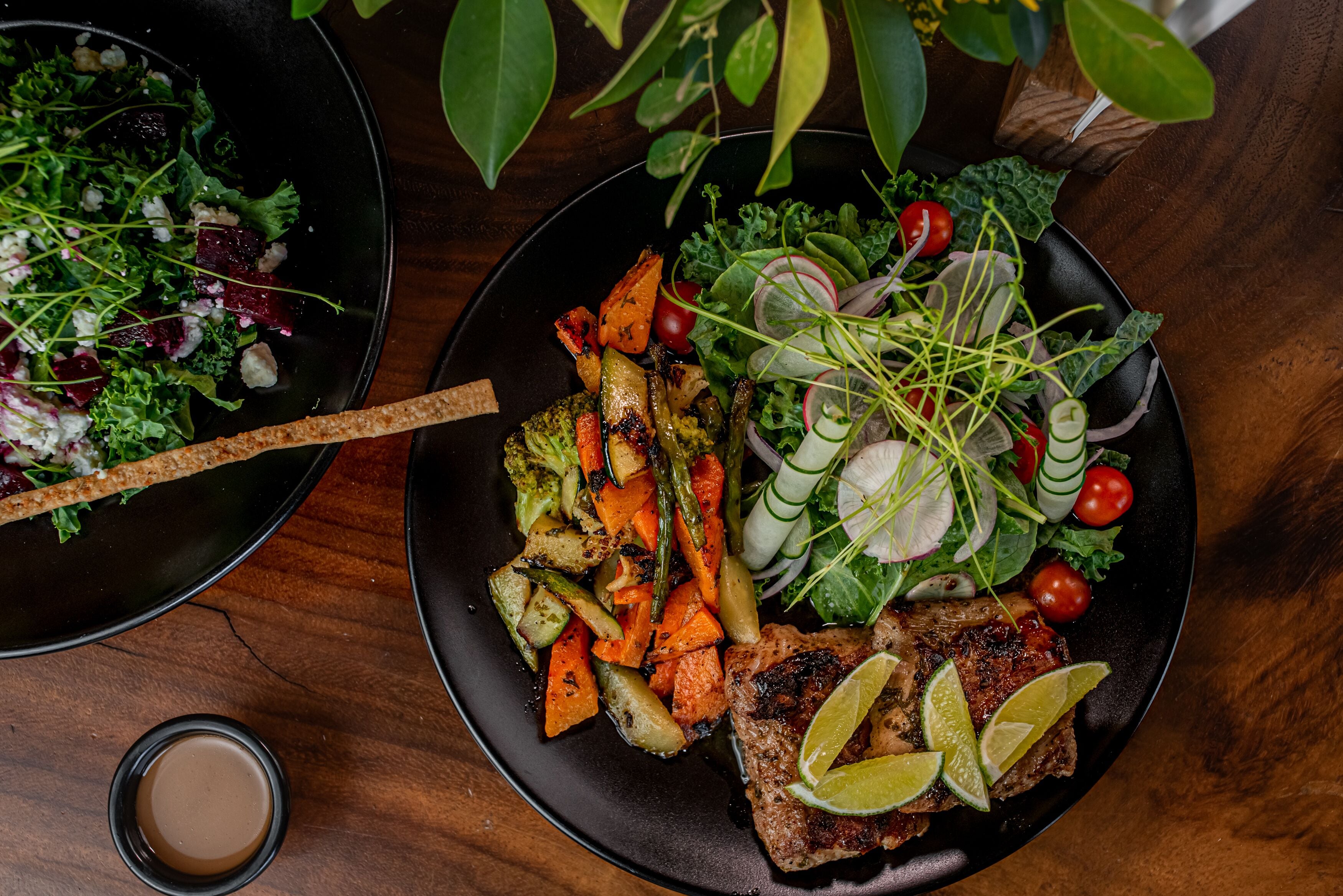 A plate of grilled vegetables and meat with lime wedges, a bowl of salad, and a small bowl of sauce on a wooden table.