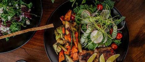 A plate of grilled vegetables and meat with lime wedges, a bowl of salad, and a small bowl of sauce on a wooden table.