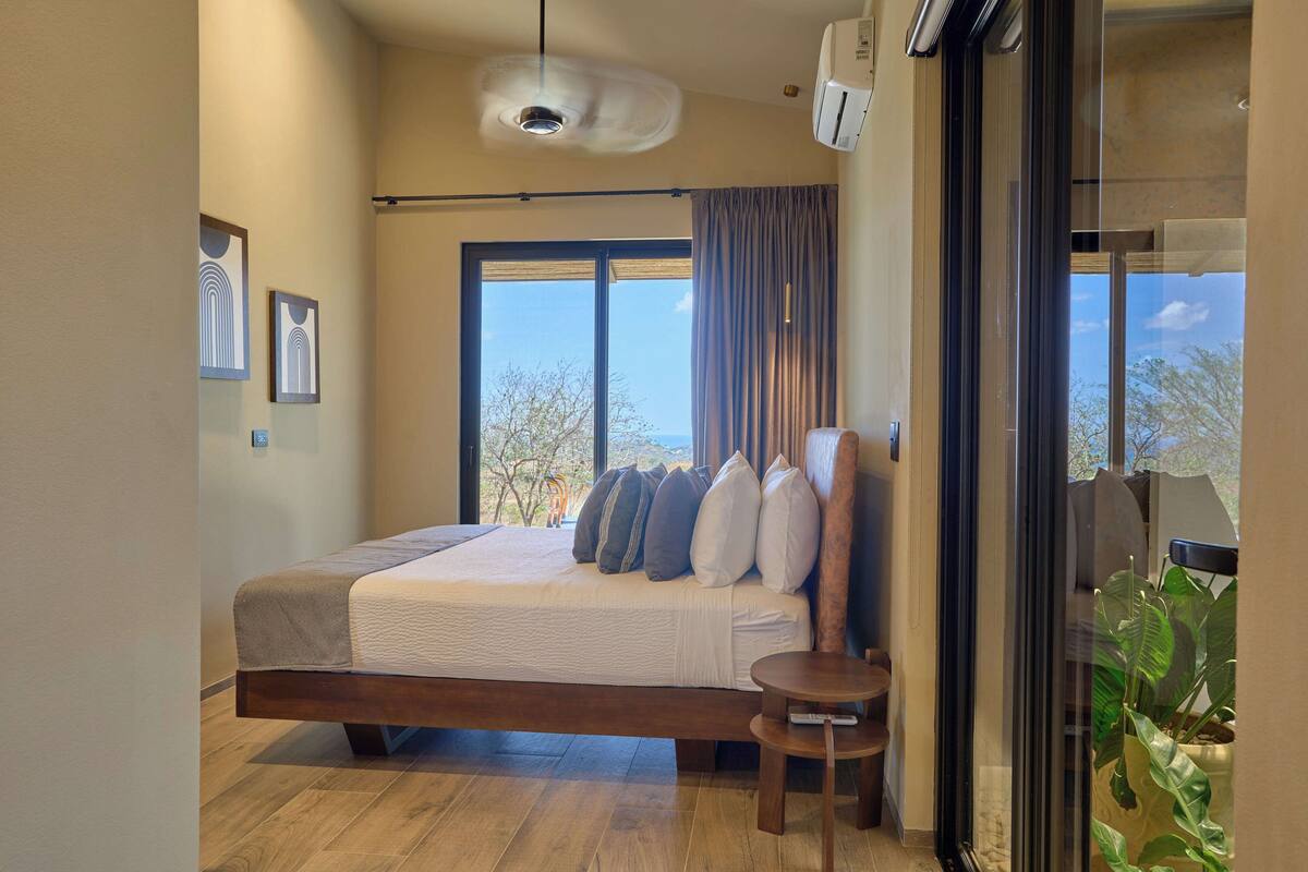A modern bedroom with a large bed, a small table, and a view of the outdoors.