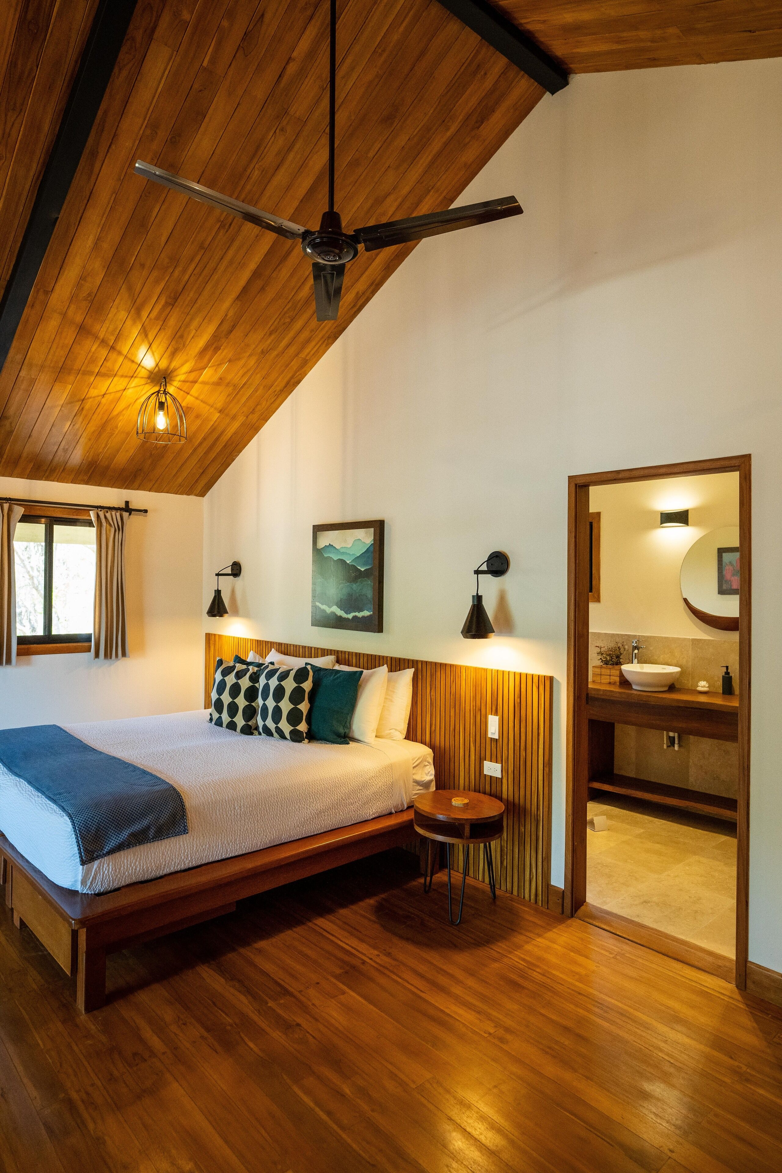 Une chambre avec un plafond en bois, un lit recouvert d’une couverture bleue, une table de chevet, une lampe fixée au mur et une salle de bain visible à travers une porte ouverte.
