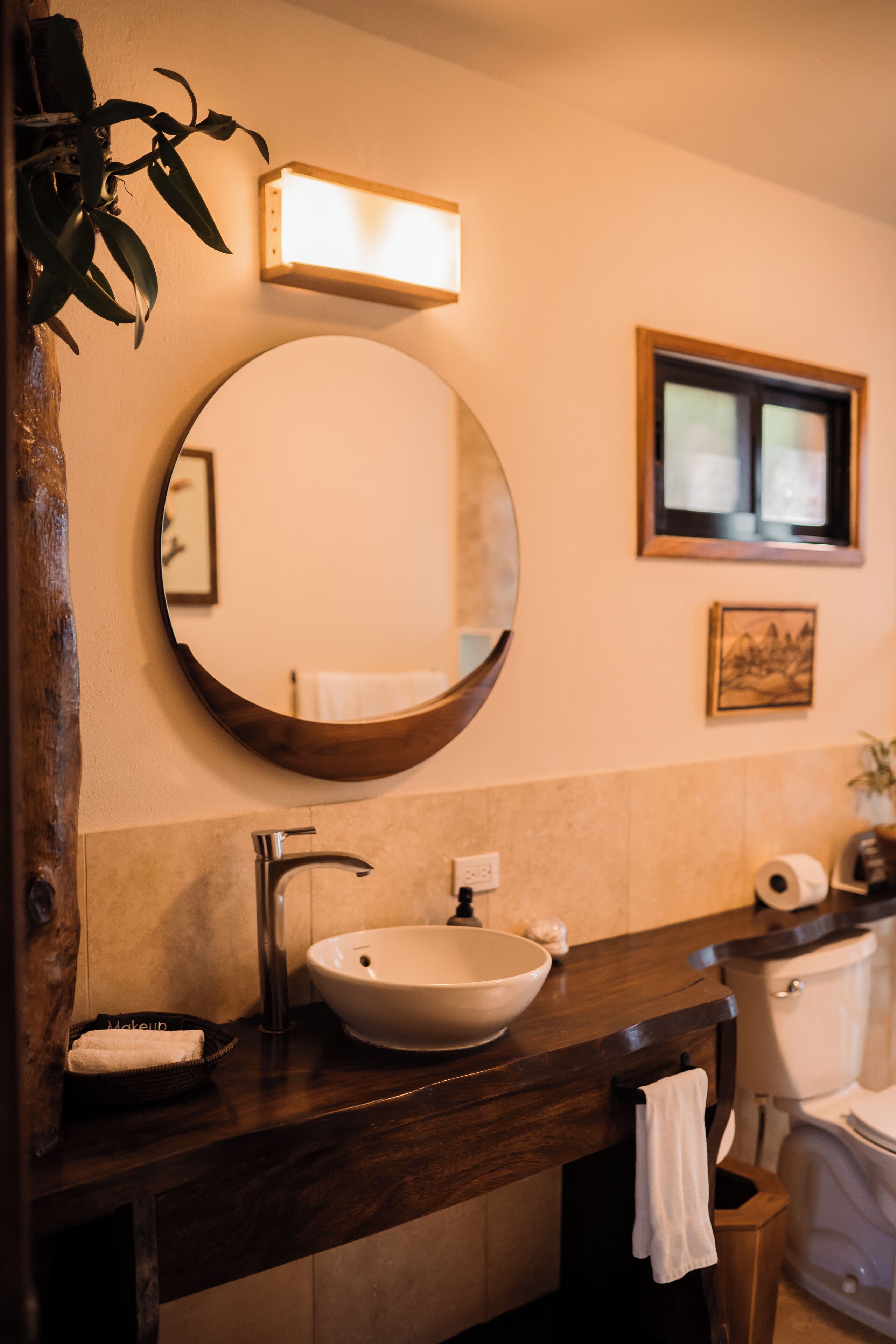 Un baño con un espejo redondo, un lavabo sobre encimera, un mueble de tocador de madera, un perchero para toallas y un inodoro.