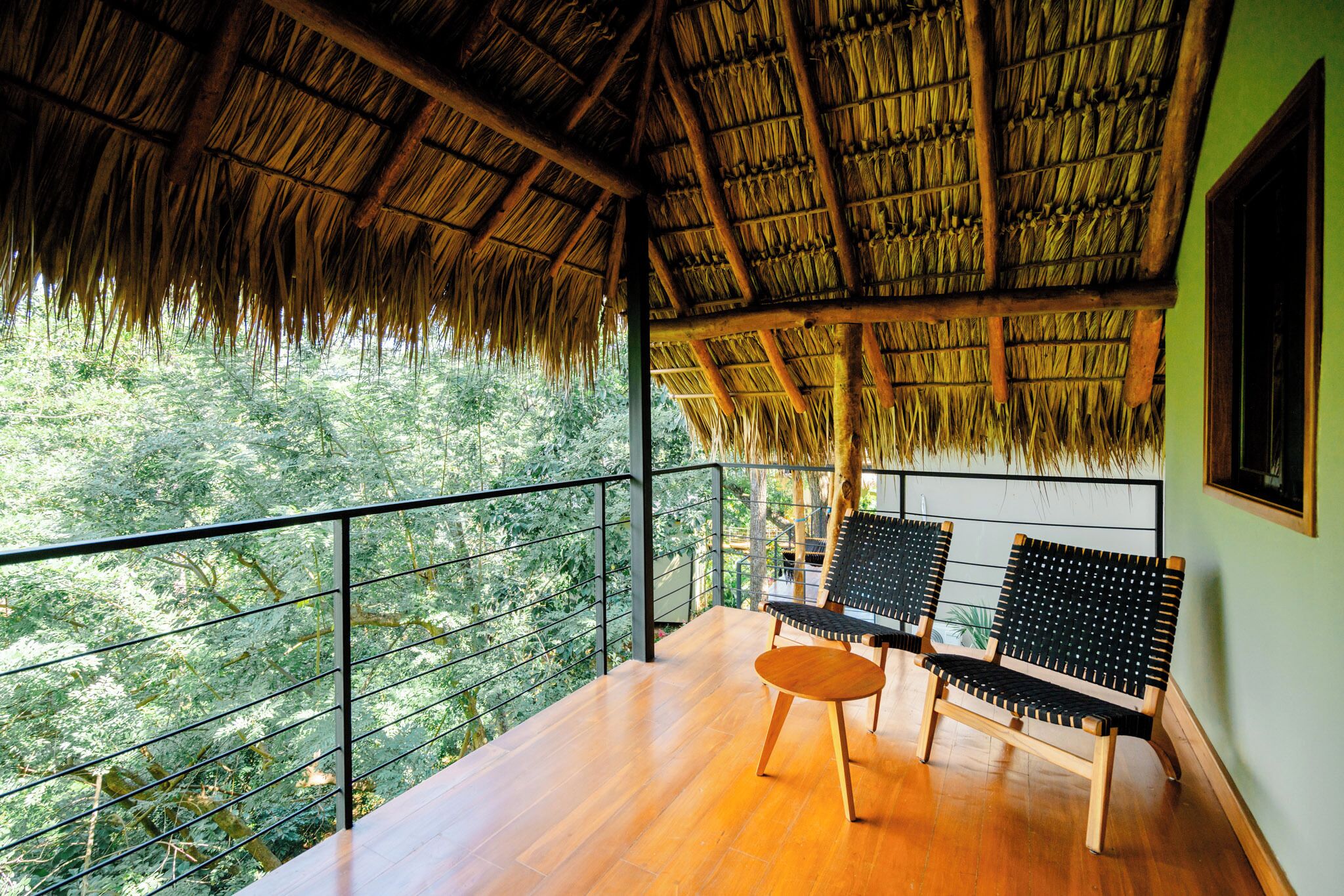 A balcony with a thatched roof, wooden flooring, and two black and wooden chairs.