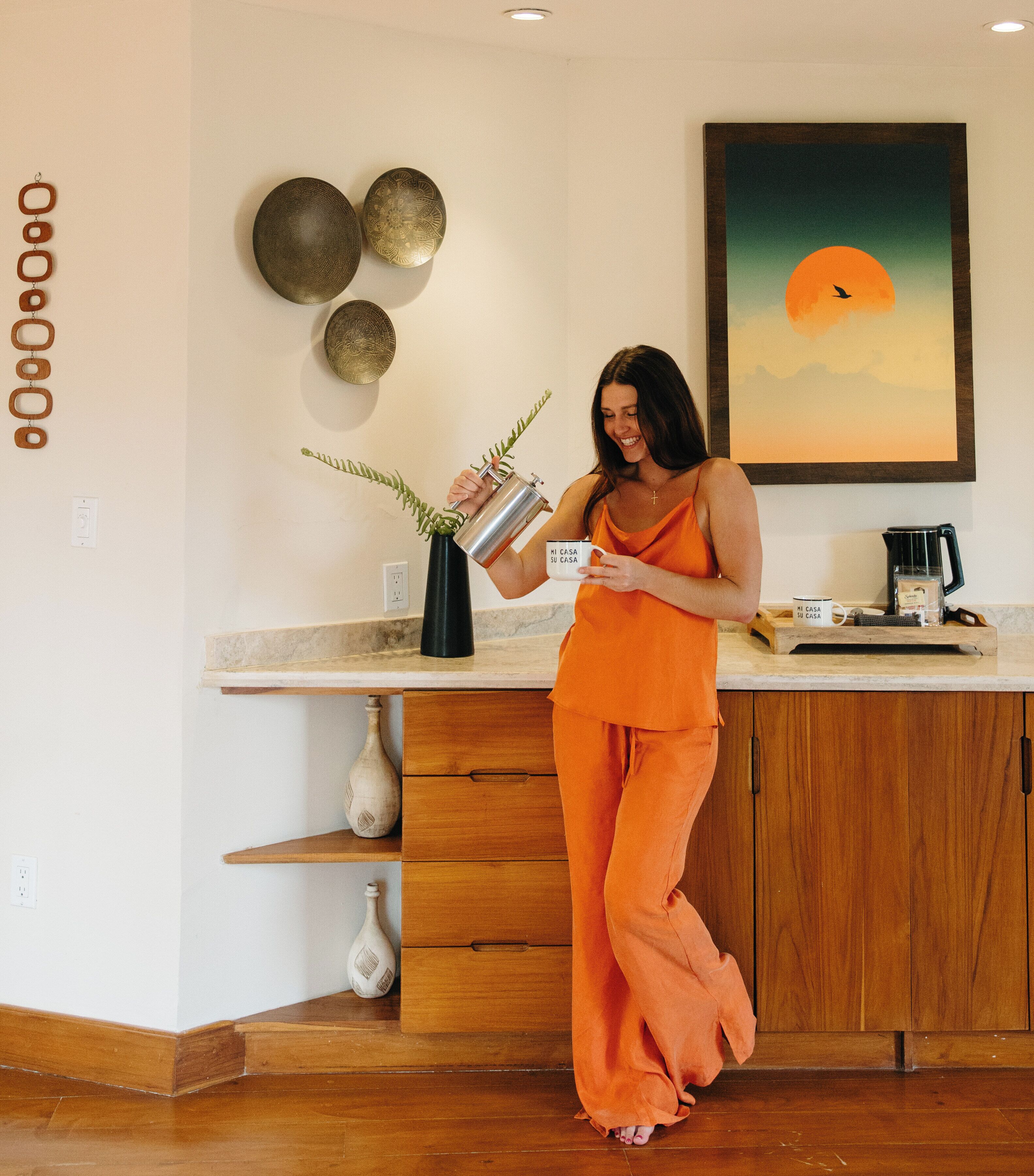 A woman in an orange outfit pouring water into a black vase on a kitchen counter with wooden cabinets and a marble countertop.