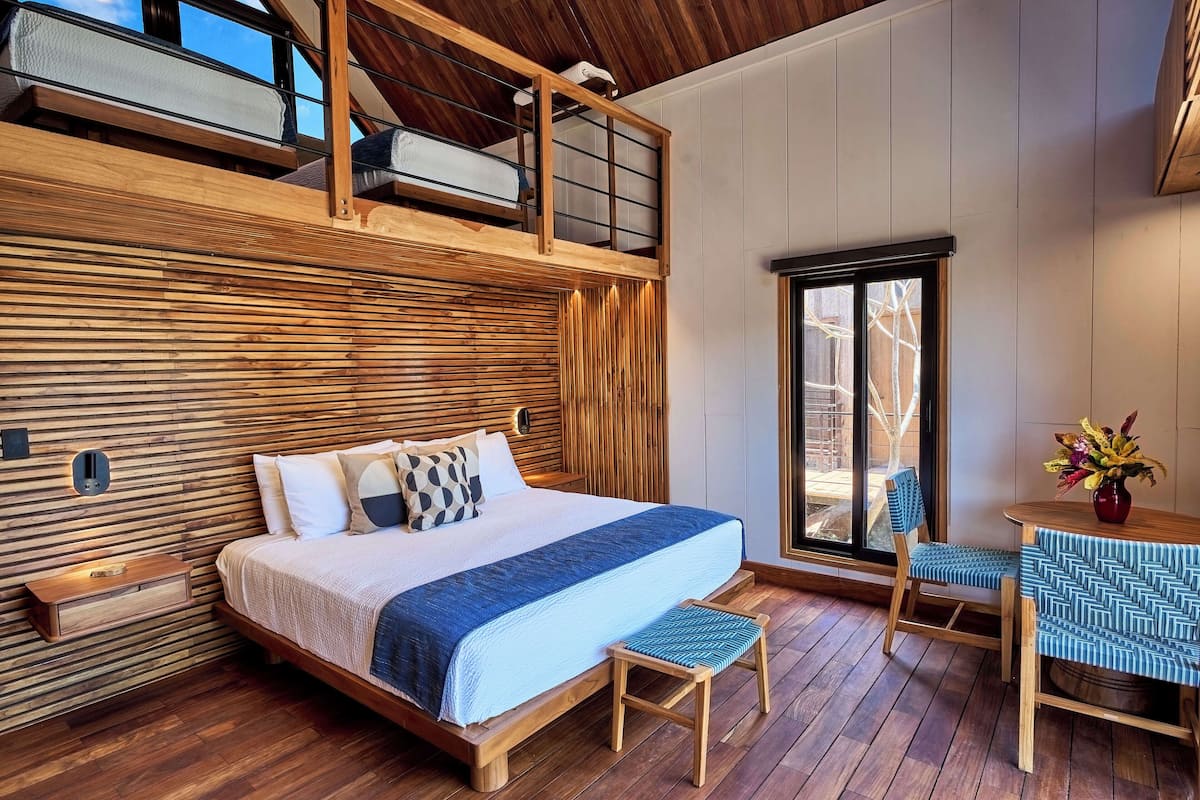 A bedroom with a bunk bed, a bedside table, a chair, a small table with a vase, and a window with a view of trees.
